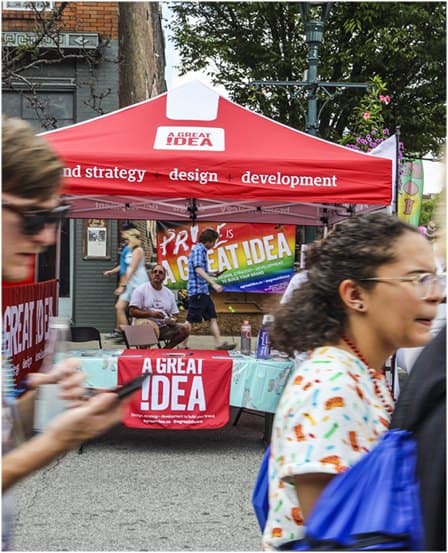 The red A Great Idea tent at a street Pride festival, with the sign 'Pride is a great idea'